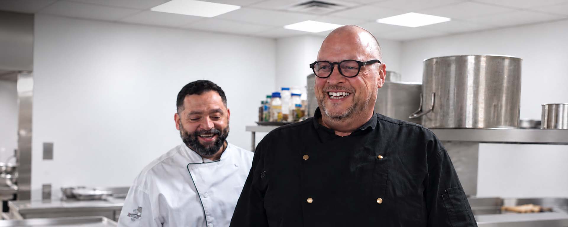 Two smiling chefs in a clean, modern commercial kitchen, showcasing professional culinary expertise and foodservice operations.