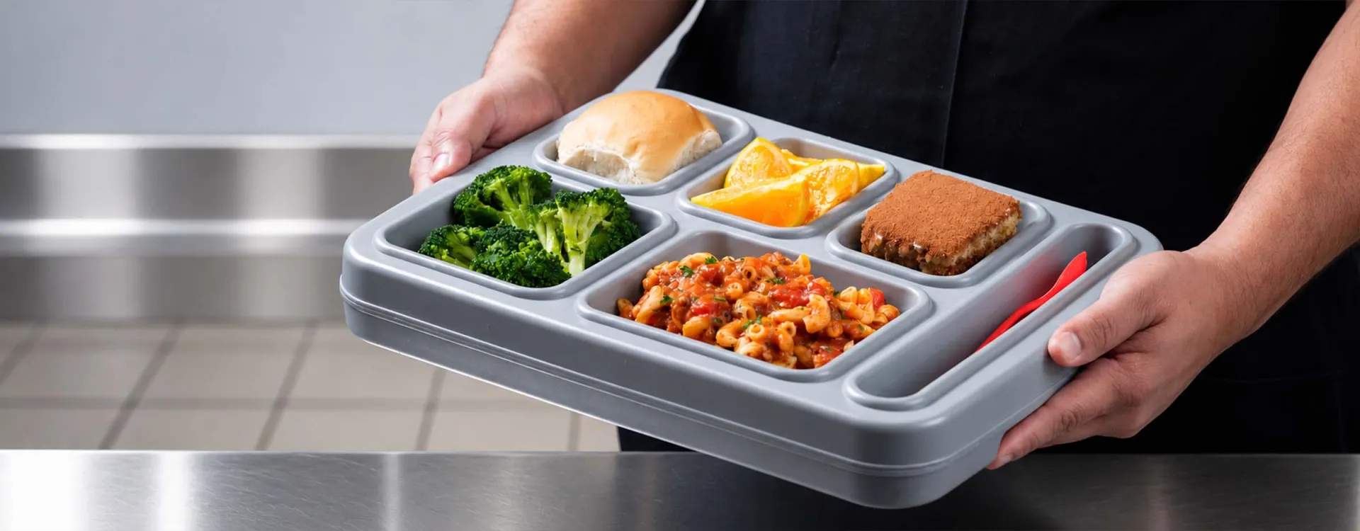 Staff member holding a balanced meal tray with pasta, steamed broccoli, fresh fruit, a dinner roll, and dessert, showcasing nutritious dining services for rehabilitation and correctional facilities.