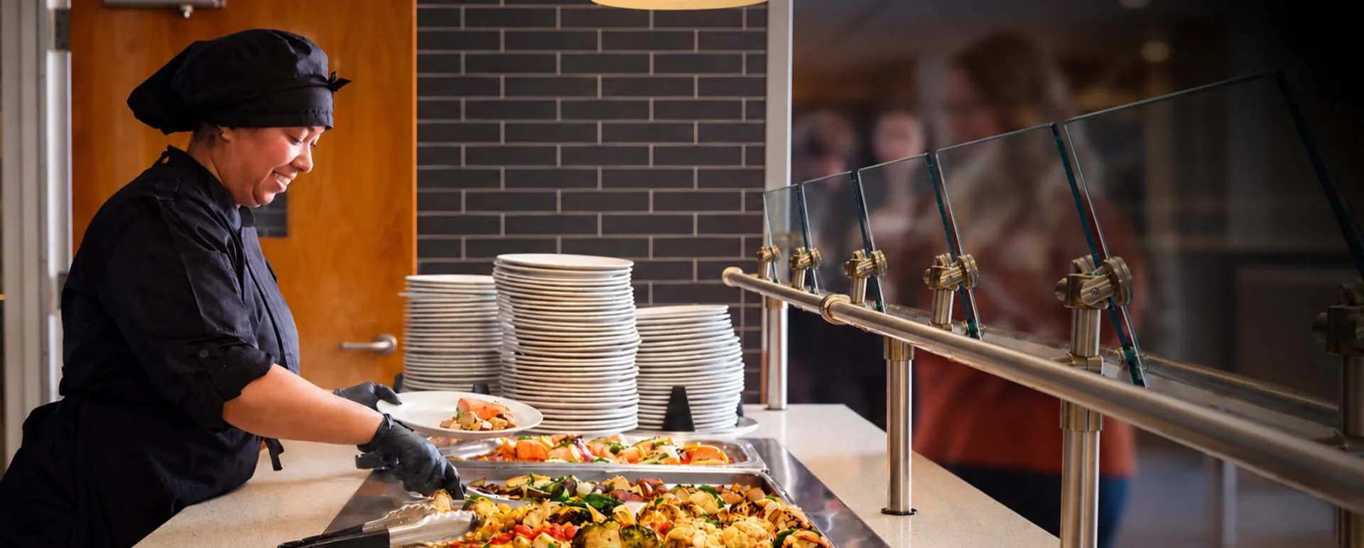  Elior North America dining team member serving a freshly prepared meal at a campus dining station, showcasing quality food and hospitality in education dining services.