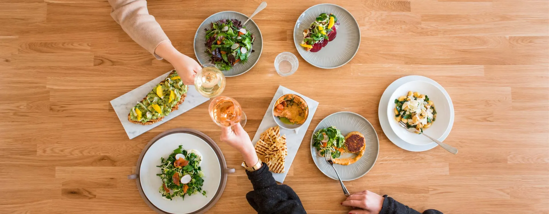 Overhead view of a shared dining experience featuring freshly prepared meals and guests raising glasses, reflecting Elior North America's commitment to culinary excellence.