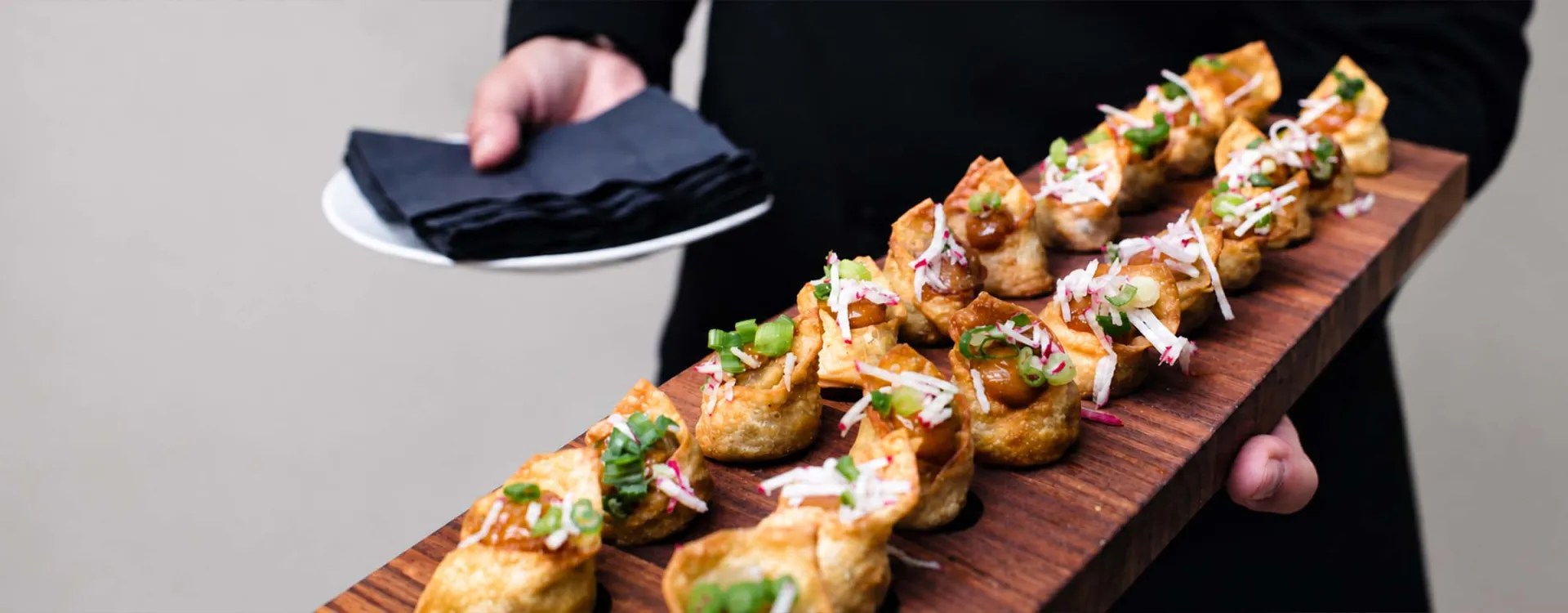 Catering server presenting freshly prepared gourmet appetizers on a wooden serving board, showcasing the culinary expertise of Elior North America.