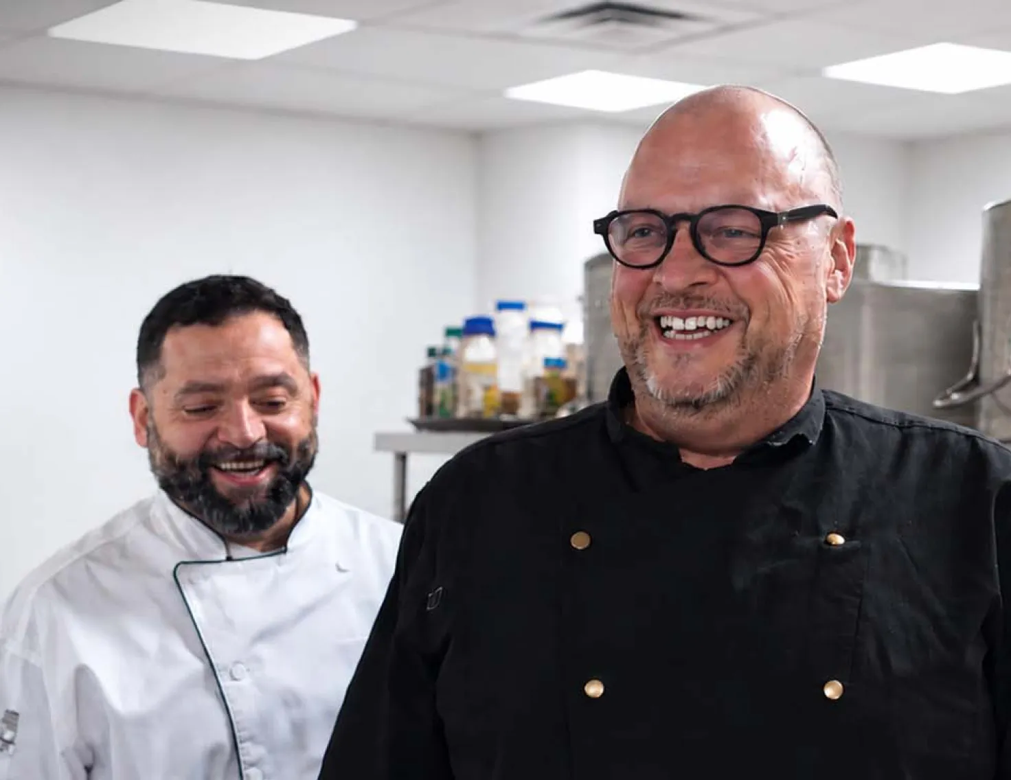 Two smiling chefs in a clean, modern commercial kitchen, showcasing professional culinary expertise and foodservice operations.