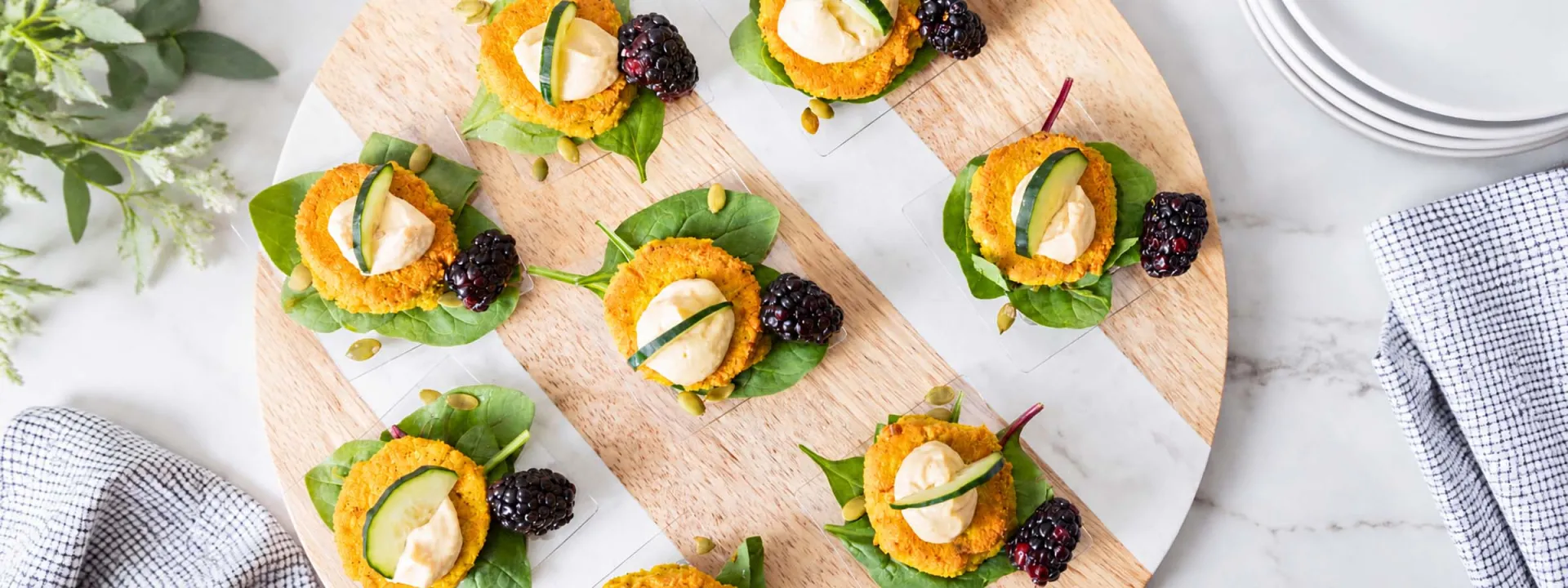 Overhead view of bite-sized appetizers topped with creamy spread, cucumber, and blackberries, arranged on a wooden serving board with fresh greens.