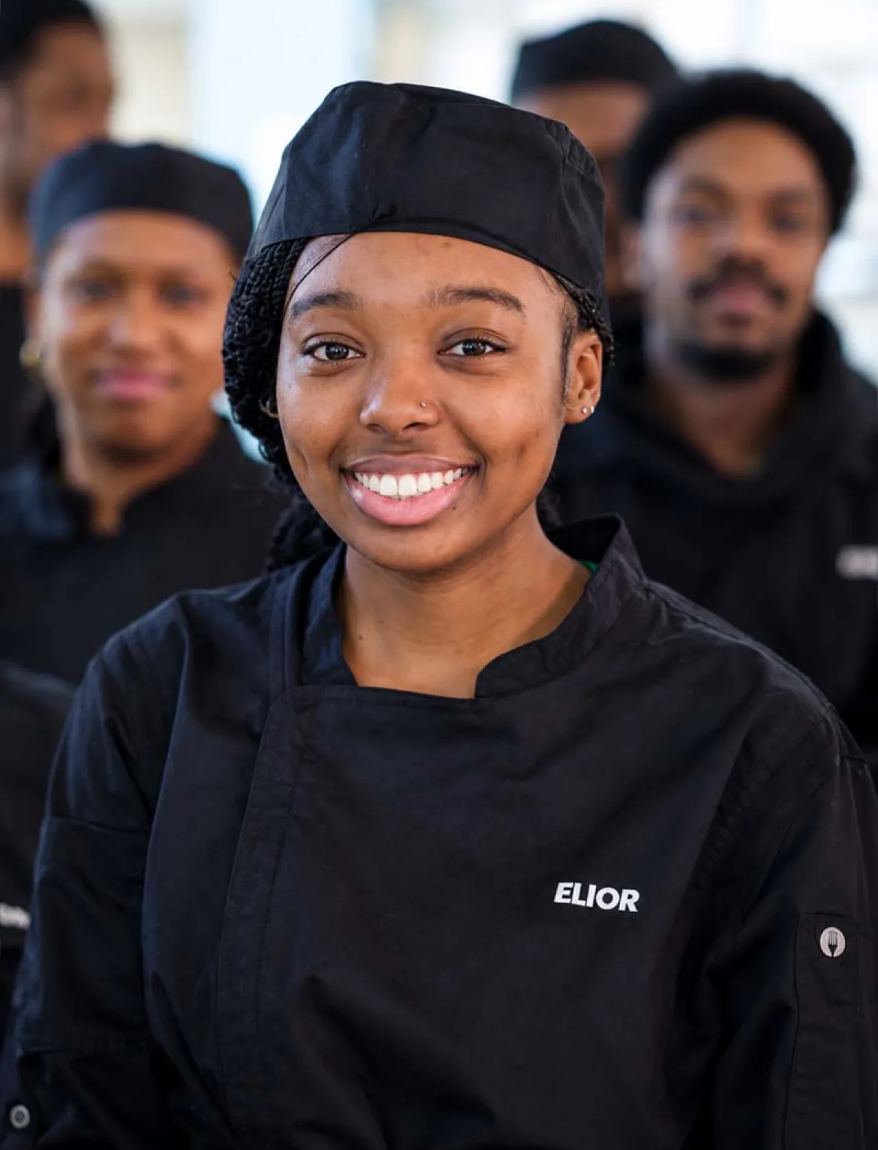 Smiling Elior North America culinary team member surrounded by colleagues, representing the people and teamwork behind our food service and hospitality operations.