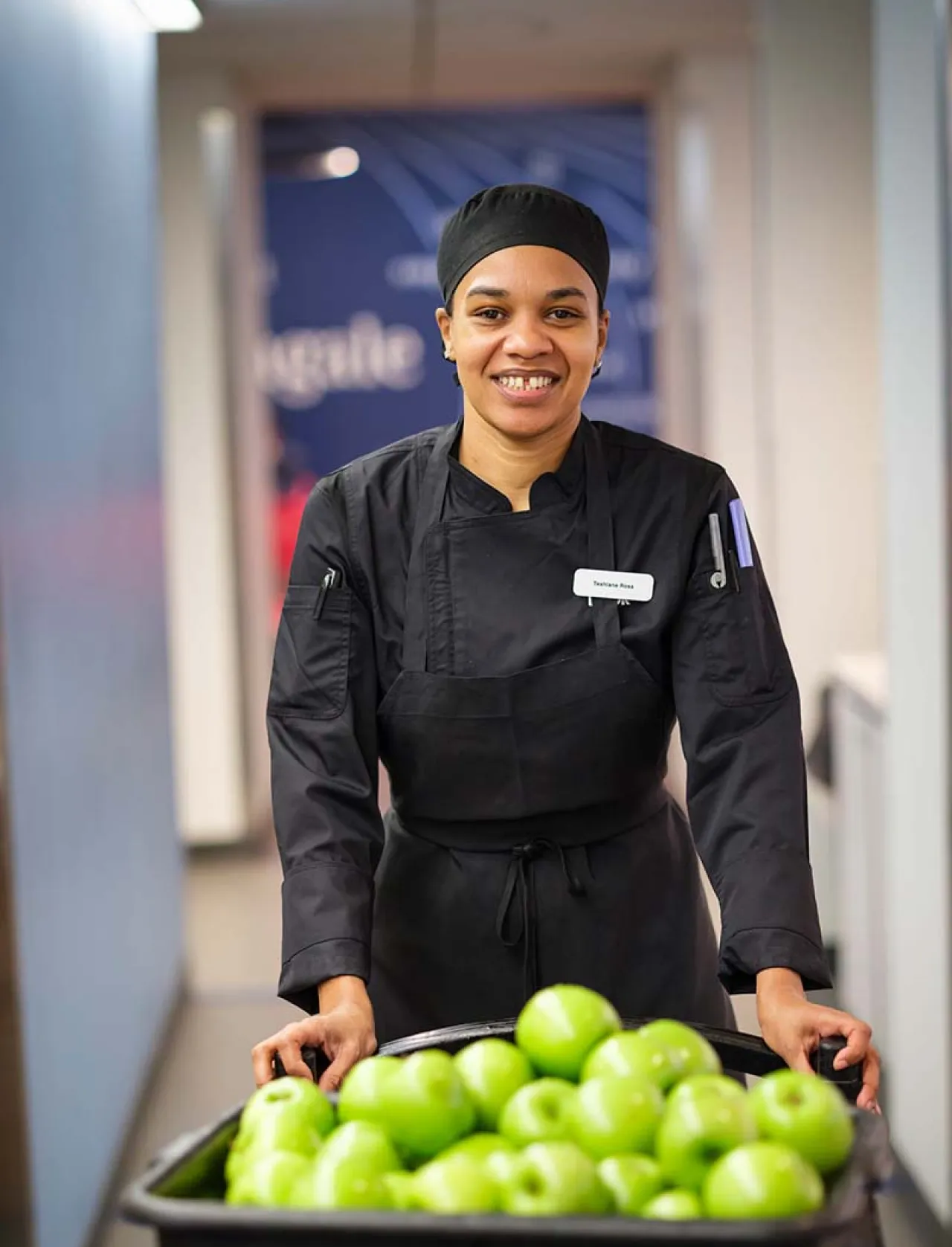 A smiling chef dressed in black pushing a cart full of green apples down a hallway.