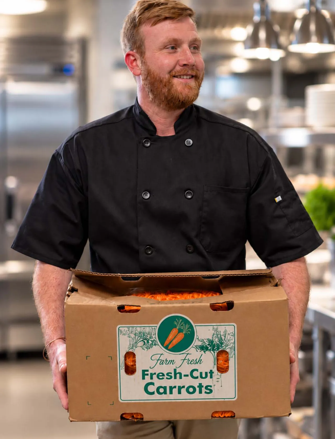 Elior North America chef holding a box of fresh carrots in a professional kitchen, highlighting responsible ingredient sourcing and sustainable foodservice practices.